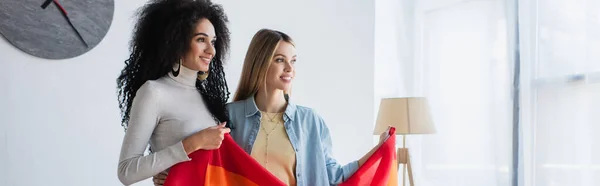 Улыбающиеся межрасовые лесбиянки, смотрящие в сторону, держа флаг, плакат — стоковое фото