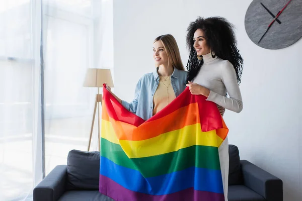 Alegre interracial lésbicas casal de pé com lgbt bandeira perto sofá em casa — Fotografia de Stock