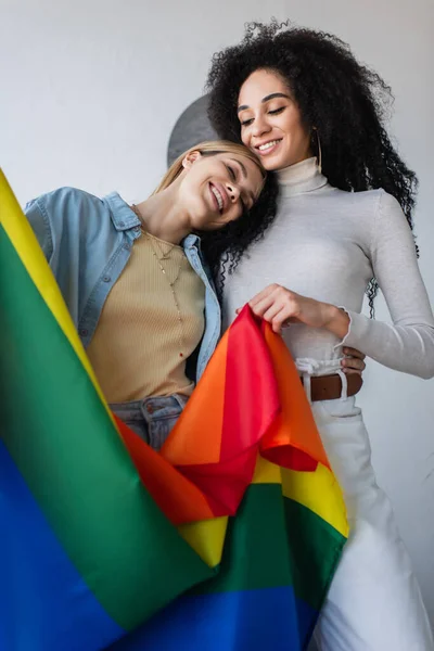 Young and happy interracial lesbian couple holding lgbt flag at home — Stock Photo