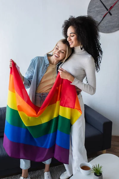 Багатонаціональні лесбійські подружки посміхаються, стоячи з lgbt прапором — стокове фото