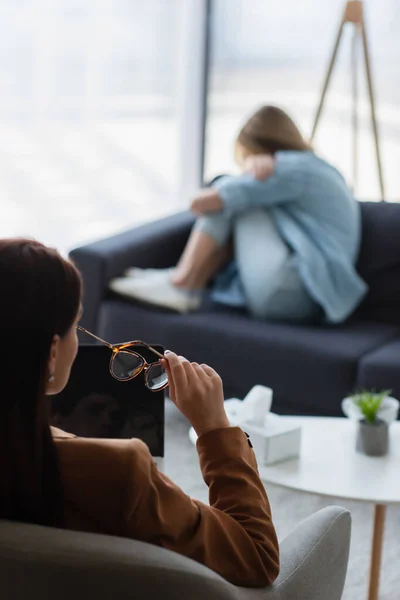 Psychologe mit Laptop und Brille in der Nähe einer depressiven Frau, die auf einem Sofa vor verschwommenem Hintergrund sitzt — Stockfoto