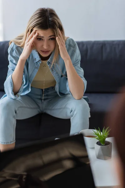 Mujer frustrada cogida de la mano cerca de la cabeza mientras está sentada cerca de psicólogo borroso - foto de stock