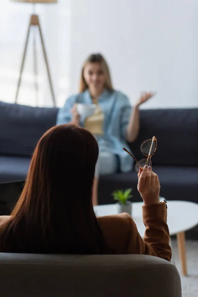 Visão traseira do psicólogo segurando óculos enquanto mulher desfocada falando em fundo desfocado — Fotografia de Stock