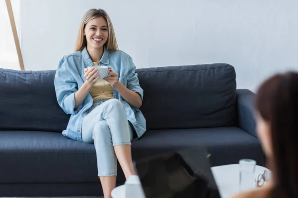 Улыбающаяся женщина, сидящая на диване с чашкой чая во время консультации с размытым психологом — стоковое фото