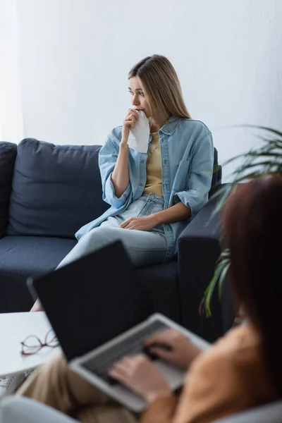 Депресивна жінка сидить на дивані біля психолога з ноутбуком на розмитому фоні — стокове фото