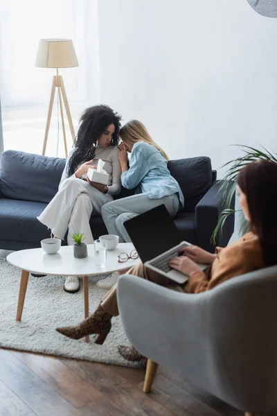 Depressed lesbian woman crying near african american girlfriend and psychologist with laptop — Stock Photo