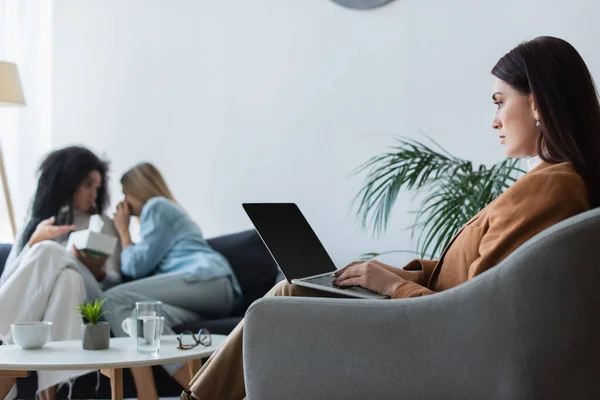 Psychologist typing on laptop with blank screen near blurred lesbian interracial couple with relationship difficulties — Stock Photo