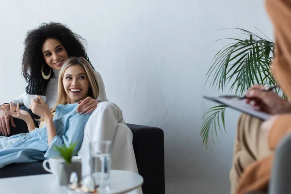 Happy interracial lesbian couple smiling during appointment with blurred psychologist — Stock Photo