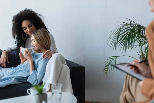 Sonriente lesbiana bebiendo té en un sofá cerca de una novia afroamericana y un psicólogo borroso escribiendo en el portapapeles - foto de stock