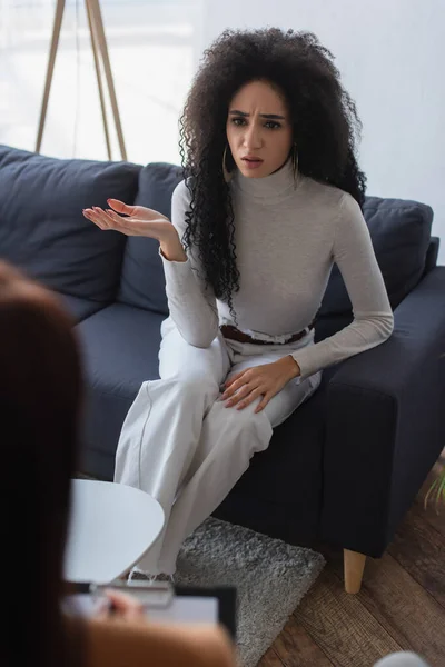 Offended african american woman sitting on couch and talking to blurred psychologist — Stock Photo