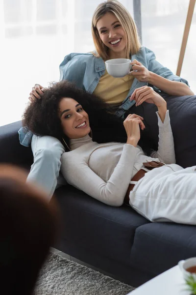 Happy woman with cup of tea near african american lesbian girlfriend and psychologist on blurred foreground — Stock Photo