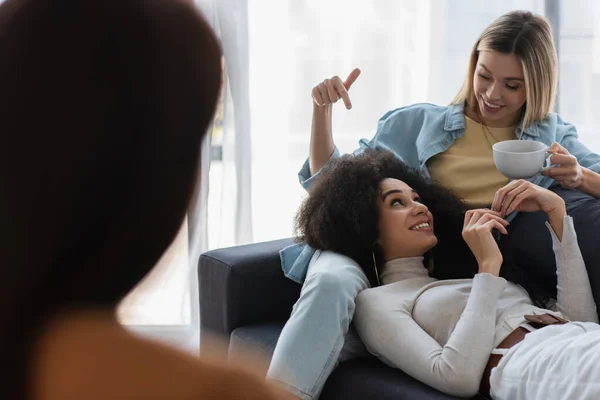 Cheerful woman with cup of tea pointing at african american girlfriend near blurred psychologist — Stock Photo