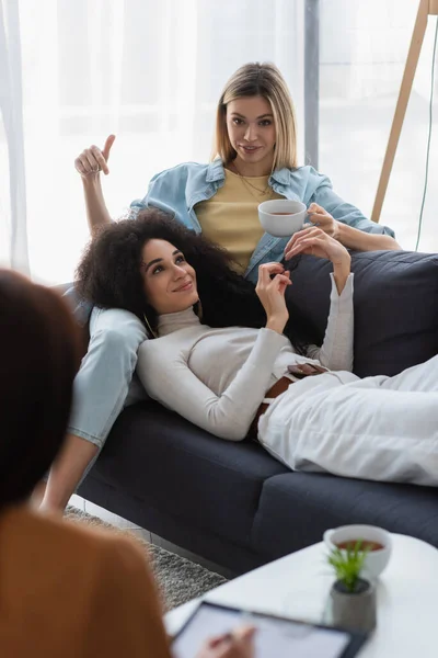 Jeune femme lesbienne avec tasse de thé pointant vers la petite amie afro-américaine pendant la consultation psychologique — Photo de stock