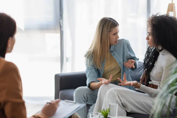 Young interracial lesbian couple quarrelling on couch near blurred psychologist writing on clipboard — Stock Photo