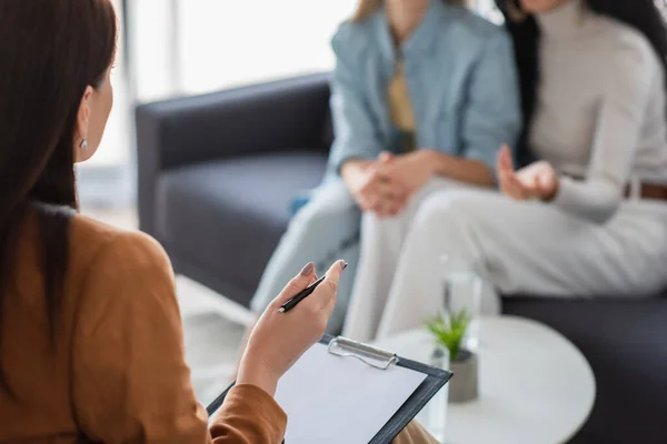 Psychologist with clipboard and pen talking to blurred lesbian couple sitting on sofa — Stock Photo