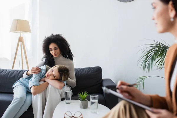 African american lesbian woman sitting with depressed girlfriend during appointment with blurred psychologist — Stock Photo