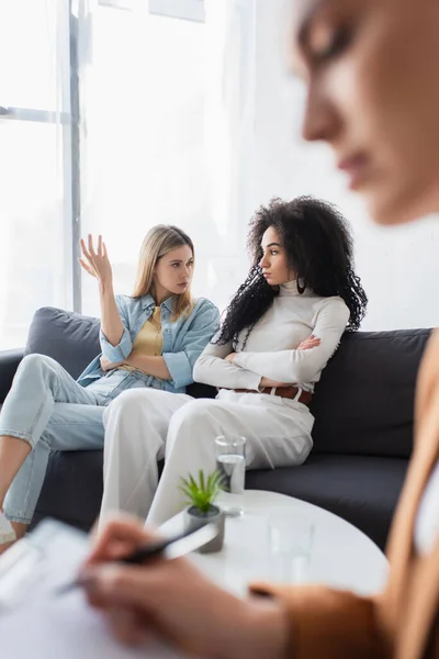 Interracial same sex couple quarreling on sofa near blurred psychologist in consulting room — Stock Photo