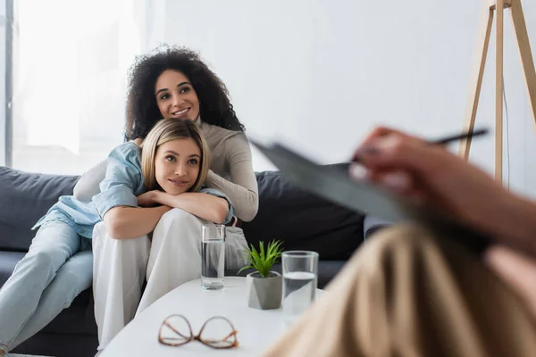 Happy interracial same sex couple listening to blurred psychologist on couch in consulting room — Stock Photo