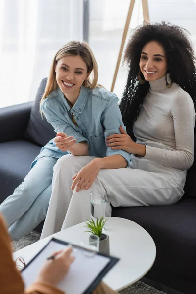 Feliz pareja de lesbianas multiétnicas sonriendo en el sofá cerca de psicólogo borrosa escritura en el portapapeles - foto de stock