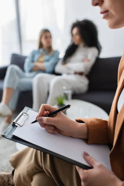 Psicólogo escribiendo en portapapeles cerca de lesbianas multiétnicas borrosas sentadas en el sofá - foto de stock