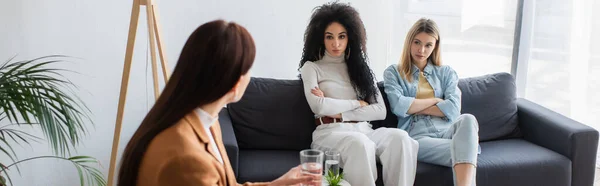 Interracial lesbians sitting with crossed arms during appointment with psychologist, banner — Stock Photo