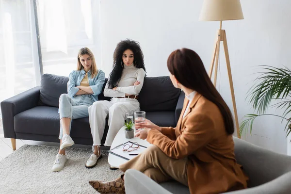 Psychologist talking to interracial lesbians sitting on couch with crossed arms — Stock Photo