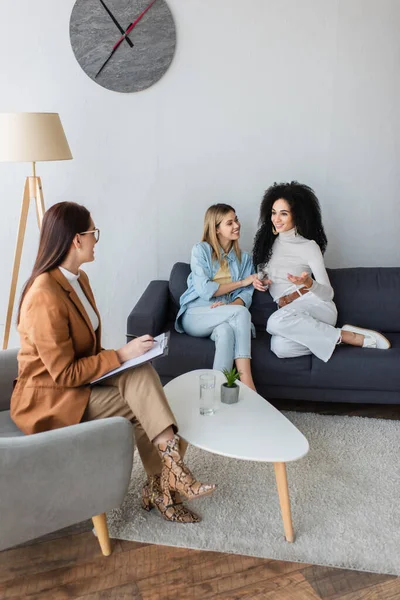 Psychologist with clipboard listening to smiling interracial lesbian couple sitting on couch in consulting room — Stock Photo
