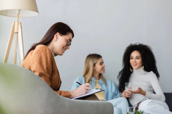 Psychologist writing on clipboard near blurred couple of interracial lesbians — Stock Photo