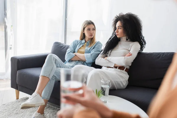 Offended interracial lesbians sitting with crossed arms on couch during psychological consultation — Stock Photo