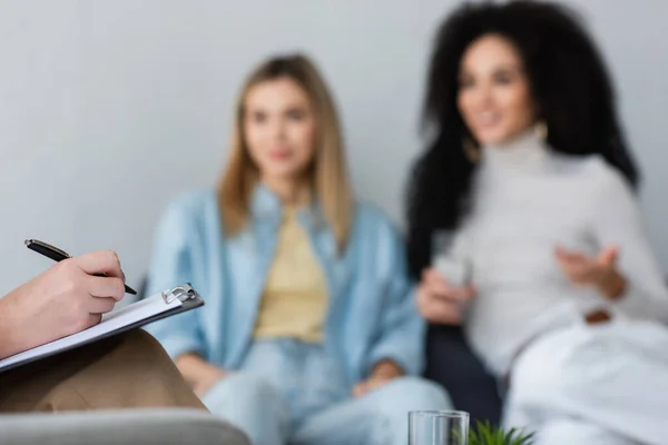 Psychologist writing on clipboard near blurred interracial same sex couple — Stock Photo