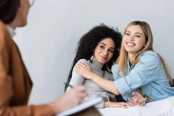 Glückliches interrassisches gleichgeschlechtliches Paar umarmt beinahe verschwommene Psychologin, die auf Klemmbrett schreibt — Stockfoto