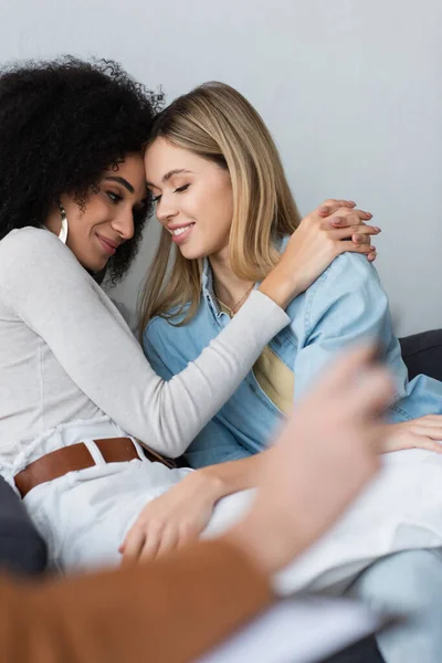 Sonriente pareja multiétnica de lesbianas abrazando cerca de psicólogo en primer plano borrosa - foto de stock