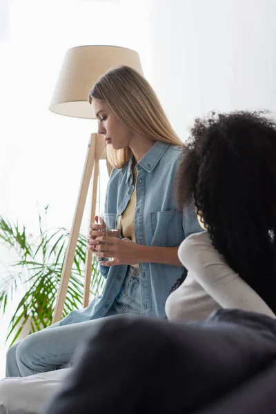 Upset lesbian woman holding glass of water near african american girlfriend — Stock Photo