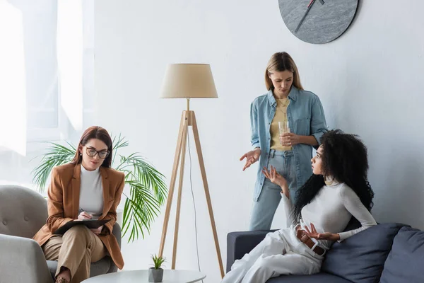 Psicólogo escribiendo en portapapeles cerca de pareja multiétnica de lesbianas hablando en la sala de consulta - foto de stock