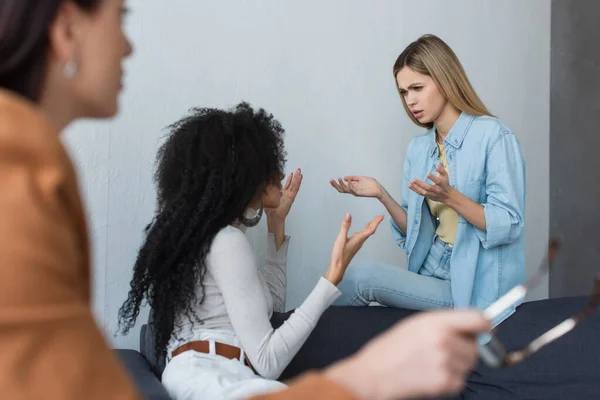 Interracial lesbian couple quarreling near blurred psychologist in consulting room — Stock Photo