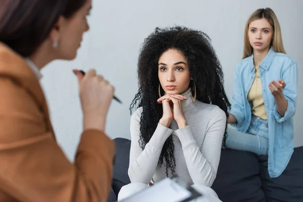 Upset couple of interracial lesbians listening to blurred psychologist during consultation — Stock Photo