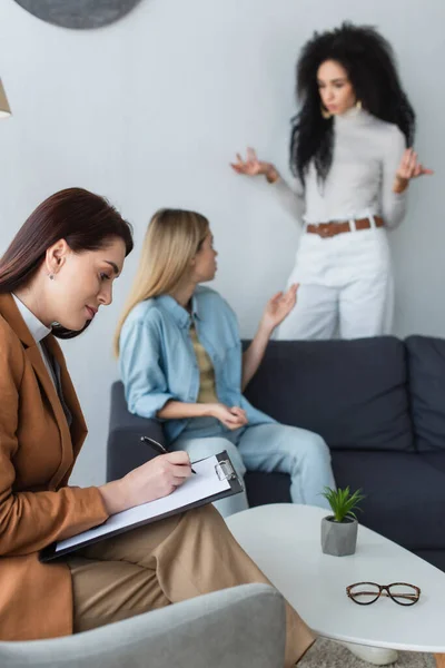 Psychologist writing on clipboard near interracial same sex couple quarreling on blurred background — Stock Photo