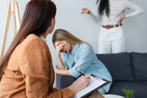 Psychologist writing on clipboard near depressed lesbian woman and her african american girlfriend — Stock Photo