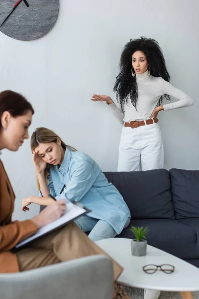 Blurred psychologist writing on clipboard near depressed lesbian woman and her girlfriend standing with hand on hip — Stock Photo