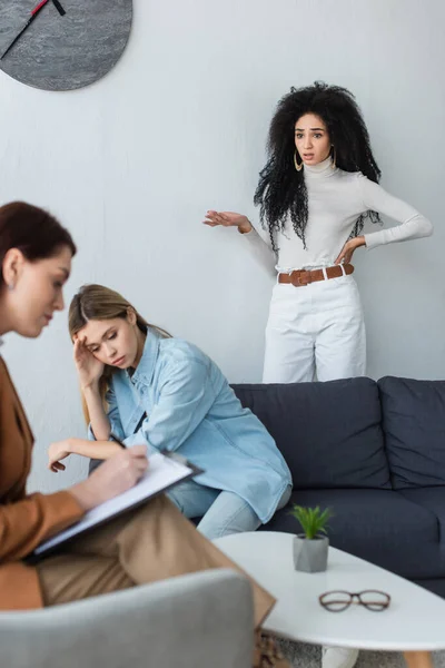 Psicóloga borrosa escribiendo en portapapeles cerca de la deprimida lesbiana y su novia de pie con la mano en la cadera - foto de stock