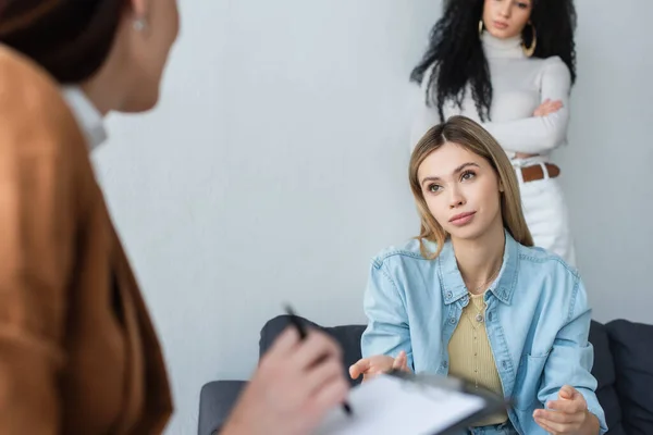 Aufgebrachte lesbische Frau sitzt neben verschwommenem Psychologen und afrikanisch-amerikanischer Freundin mit verschränkten Armen — Stockfoto