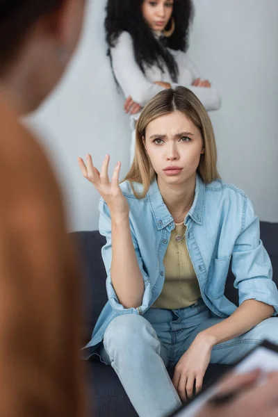 Upset lesbian woman talking to blurred psychologist near african american girlfriend standing with crossed arms — Stock Photo