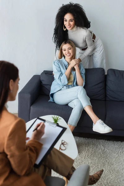 Blurred psychologist writing on clipboard near happy multiethnic same sex couple — Stock Photo