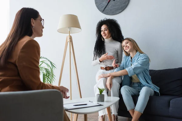 African american woman pointing at lesbian girlfriend while talking during consultation with blurred psychologist — Stock Photo