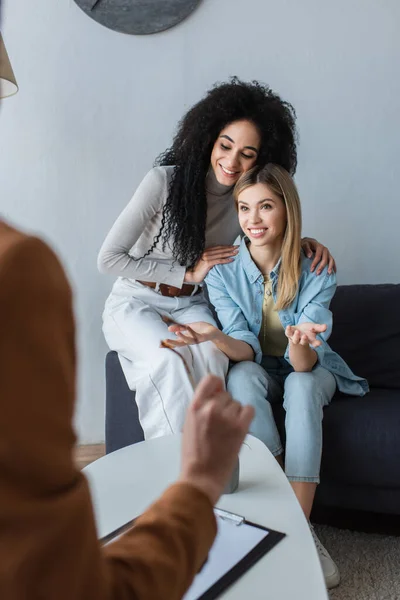 Mulher sorridente gesticulando enquanto conversava com psicólogo turvo perto da namorada afro-americana — Fotografia de Stock