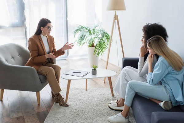 Psicólogo positivo gesticulando mientras habla con pareja interracial de lesbianas en la sala de consulta - foto de stock