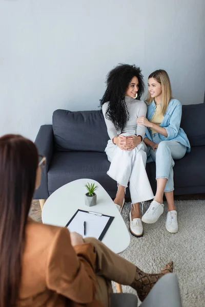 Smiling interracial lesbian couple looking at each other while sitting on couch during appointment with psychologist — Stock Photo