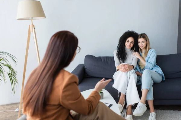 Psicóloga borrosa señalando con la mano mientras habla con lesbianas multiétnicas felices sentadas en el sofá - foto de stock