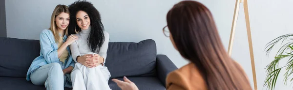 Blurred psychologist talking to smiling multiethnic lesbian couple in consulting room, banner — Stock Photo