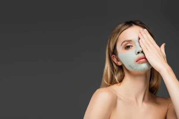 Young woman with naked shoulders and clay mask obscuring face with hand isolated on grey — Stock Photo